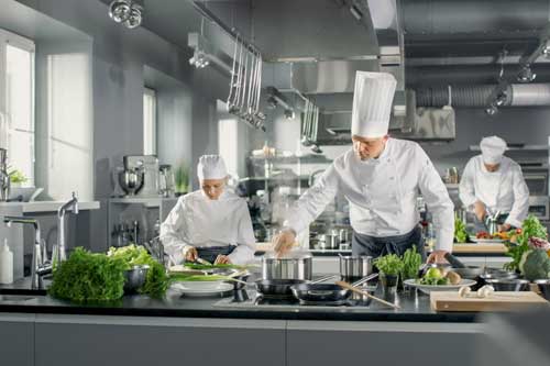 Küchenmeister Ausbildung IHK | Der berühmte Küchenchef arbeitet mit seiner Hilfe in einer großen Küche. Die Küche ist voll von Essen, Gemüse und Kochgeschirr.
