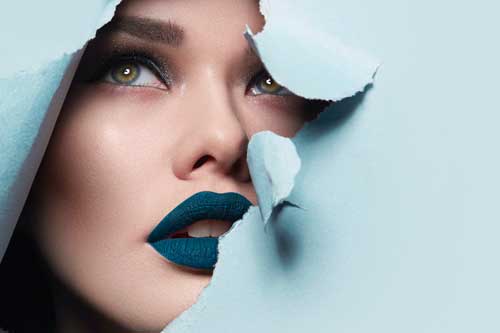 Fernstudium Visagist/in  | das Gesicht eines jungen schönen Mädchen mit einem hellen Make-up und puffblauen Lippen Peers in ein Loch in blauem Papier.